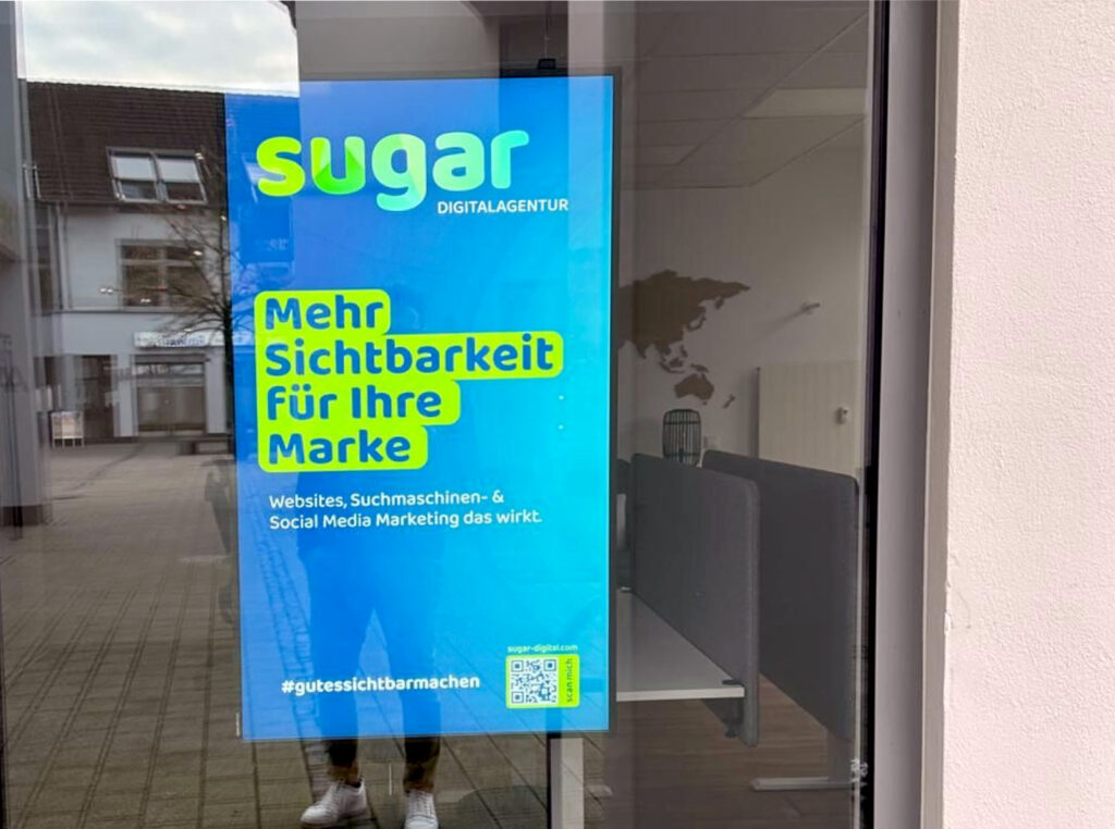 Werbefläche im Fenster mit einem auffälligen Plakat der 'Sugar Digitalagentur', das für mehr Sichtbarkeit von Marken durch Websites, Suchmaschinen- und Social Media Marketing wirbt, ergänzt durch einen QR-Code für weitere Informationen.